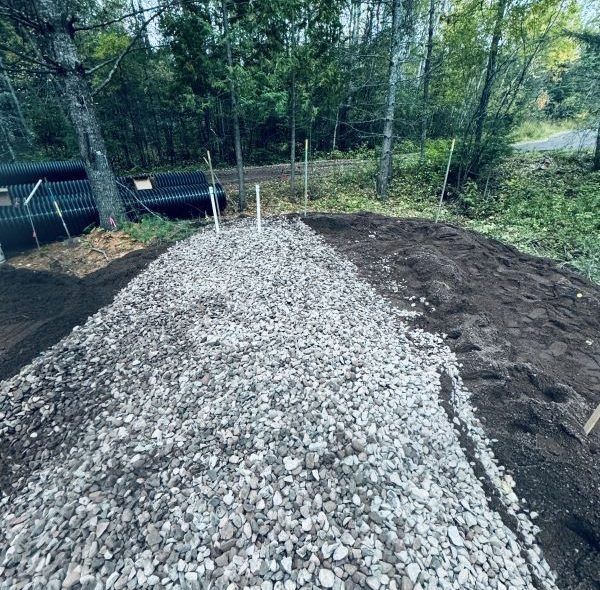 Pile of gravel and dirt for a construction project near trees and black pipes.