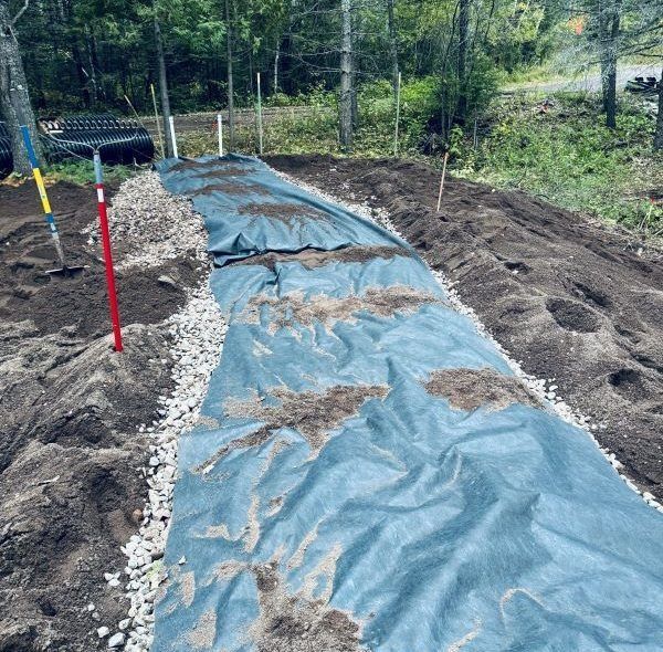 Blue landscape fabric laid in garden bed, bordered with gravel. Tools standing nearby.