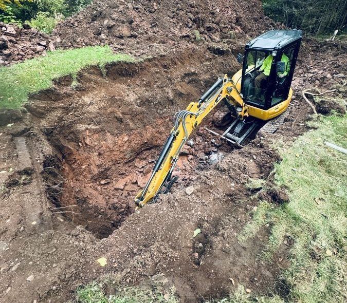 Yellow excavator digging a rectangular hole in brown earth; operator visible. Green grass surrounds.