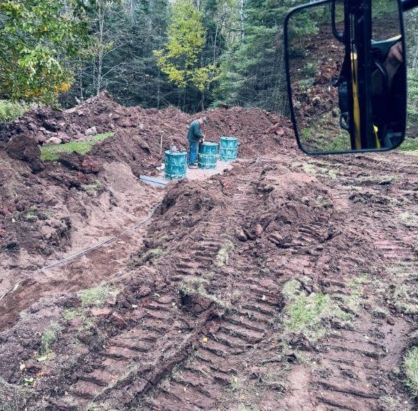 Person near blue barrels in a dirt excavation. Forest background, vehicle mirror on the right.