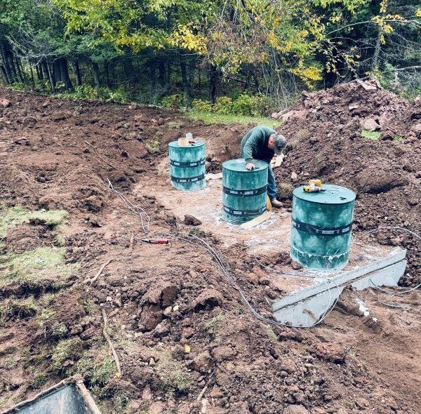 Construction site: man installing green septic tanks in dirt. Trees in background.