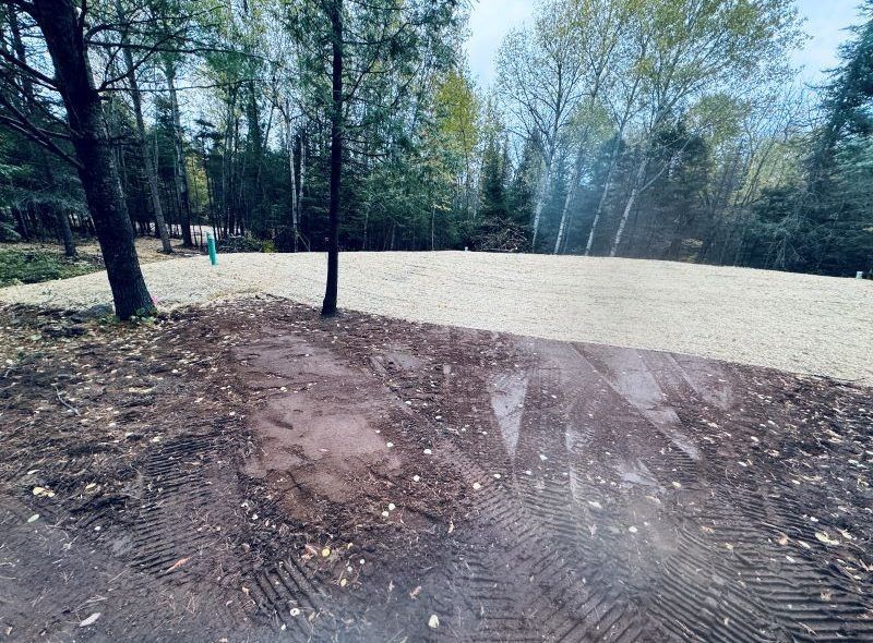 Cleared land with gravel base, tire tracks in foreground, trees in background.