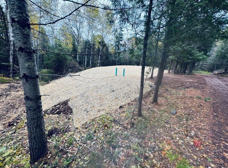 Gravel bed in a wooded area, likely for construction. Blue pipes protrude. Brown ground, green trees.