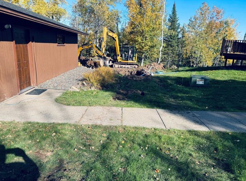 Construction site: Backhoe digging near a brown building and sidewalk on a green lawn with trees.