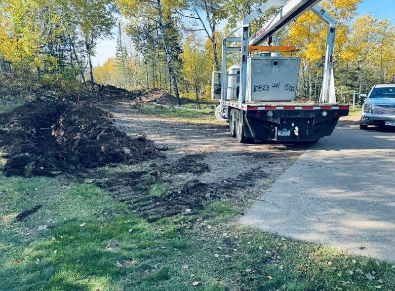 Flatbed truck carrying concrete tank near a construction site, with a muddy path and parked vehicles.