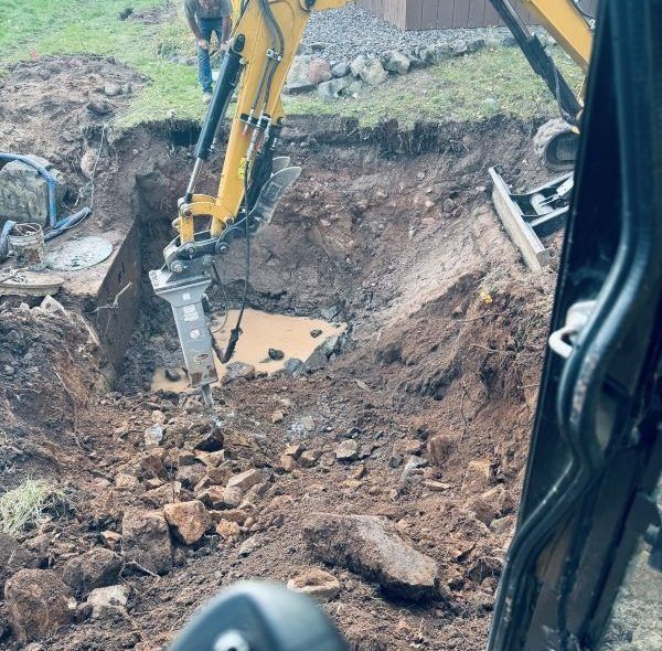Yellow excavator breaking apart concrete in a deep excavation, brown earth, bright sunlight.