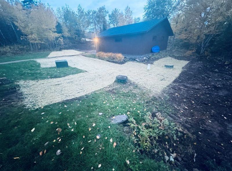 A gravel driveway leads to a small brown building surrounded by green grass and trees.