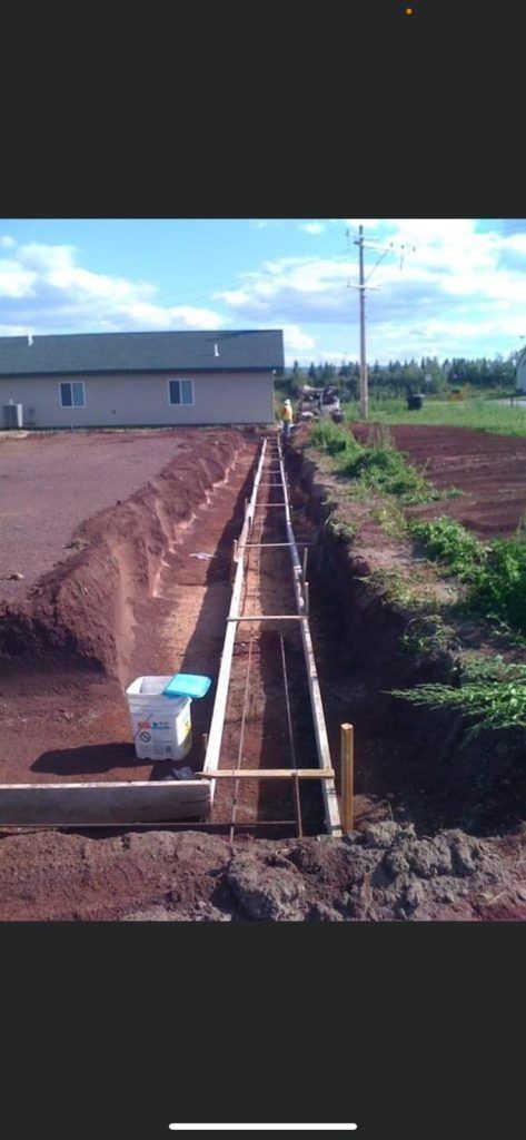 Construction site, trench with wooden forms, a building, and a bucket. Red soil, blue sky, and some greenery.