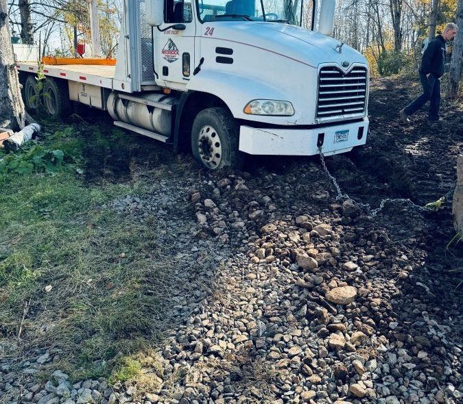 White tow truck stuck in mud and rocks, pulling a cable, man walking nearby.