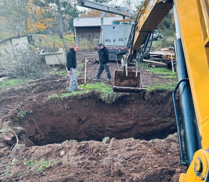 Two men watch an excavator dig a large hole in a yard.