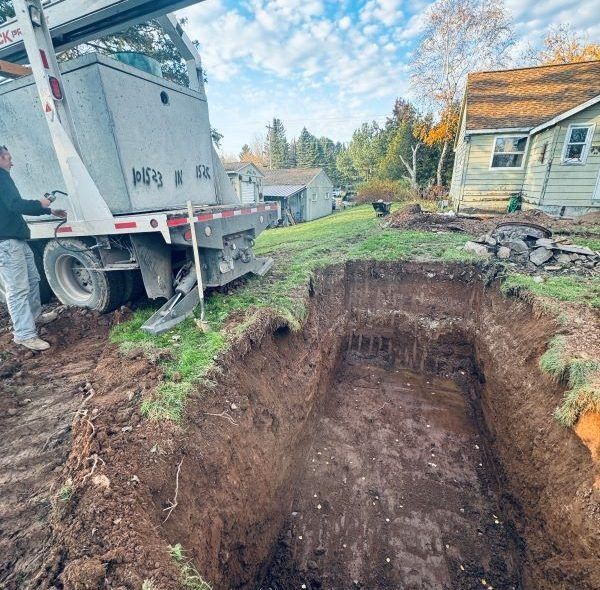 A man and a crane installing a concrete tank into an excavated hole next to a house.