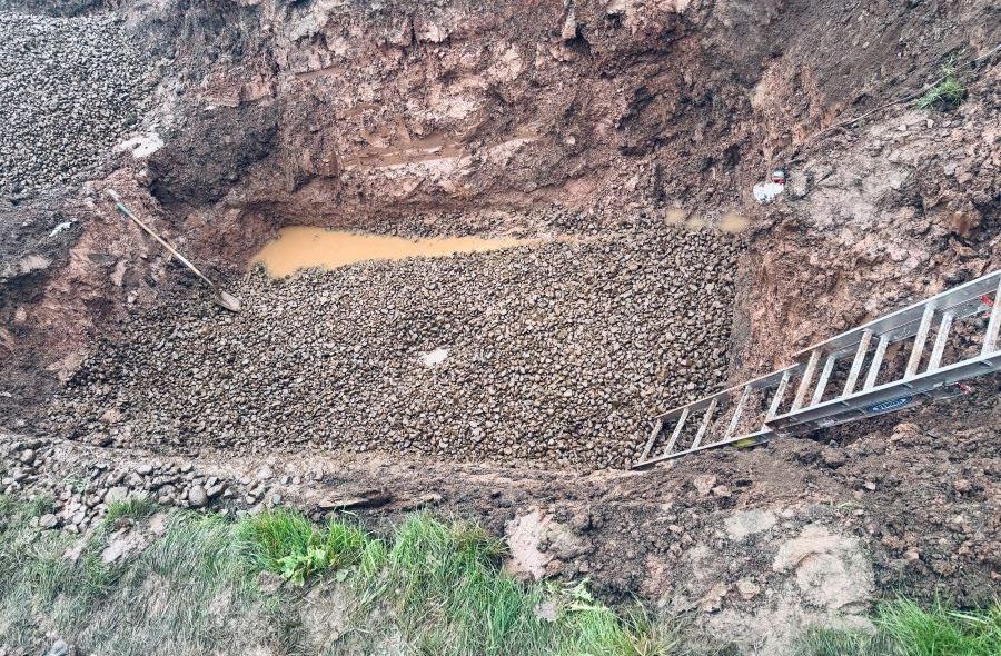 A cut bank with a pile of rocks at the base, a ladder, and brown soil and grass.
