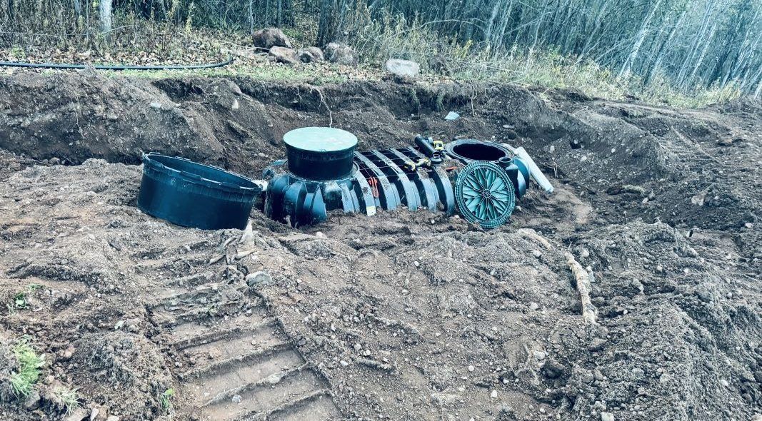 Septic tank components in a dug-out trench surrounded by dirt and vegetation.