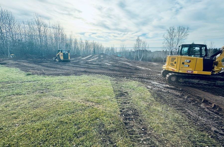 Construction site with yellow excavator and skid steer on muddy ground, clearing land near trees.