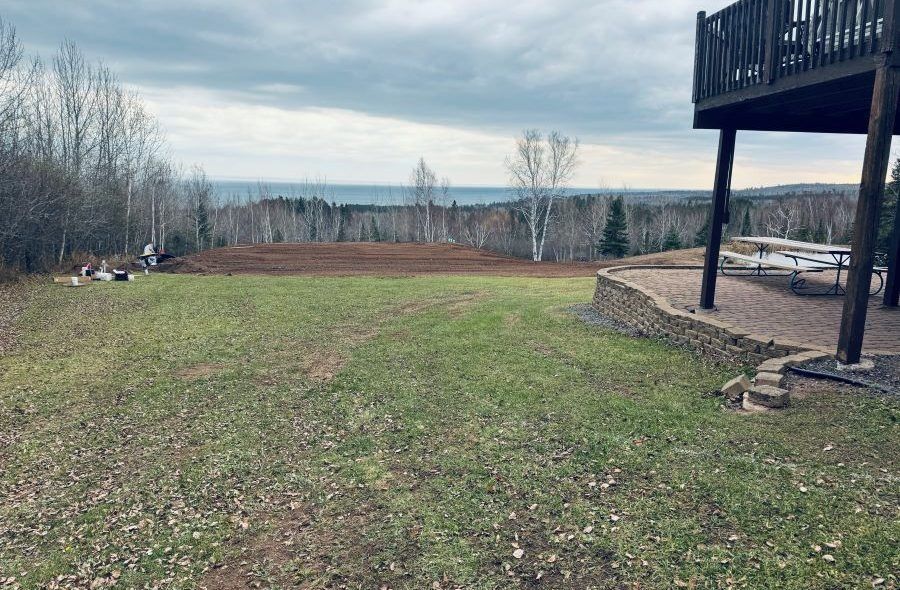 Green lawn and cleared dirt patch overlook a lake under a cloudy sky, beside a wooden deck.