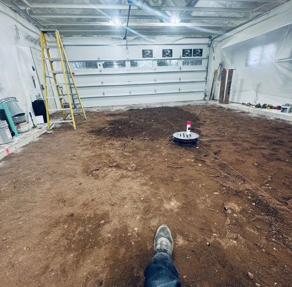 Person in jeans and sneakers standing in a dirt-floored garage; ladder and garage door visible.