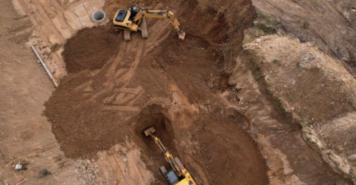 Two yellow excavators digging in a brown dirt construction site. Aerial view.