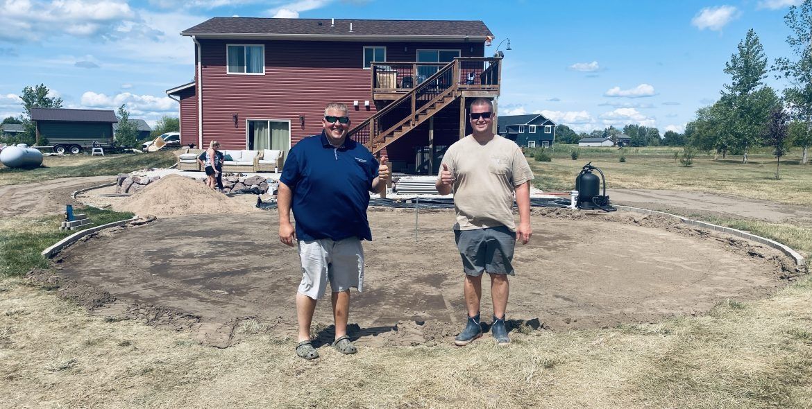 Two men stand in a dirt yard in front of a red house; construction site.