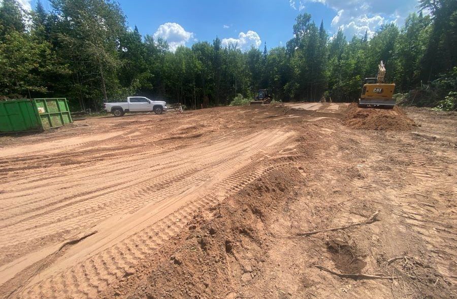 Clearing site: truck, excavator, green bin on a dirt area, trees in background, sunny day.
