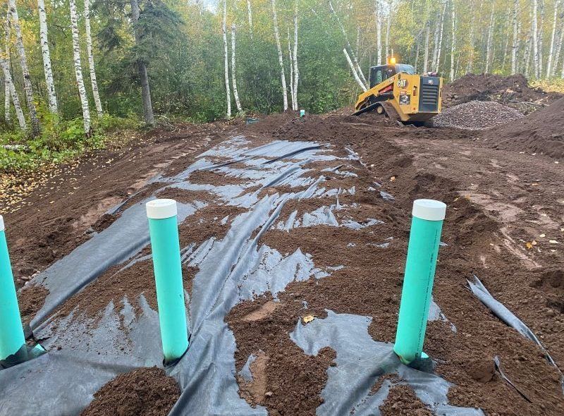 Construction site: Yellow bulldozer, black fabric, green pipes, and dirt with trees in the background.
