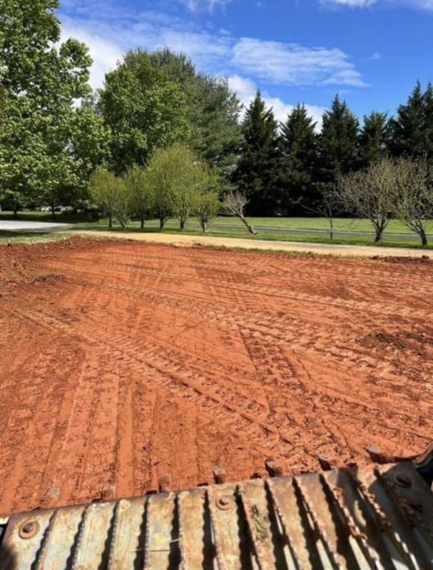 Red dirt and tire tracks fill a cleared construction site. Green trees and blue sky in the background.