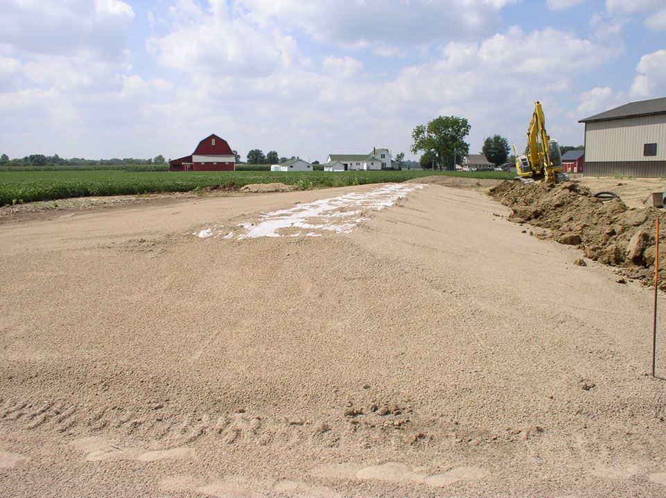Road construction with gravel base, farmland in background.