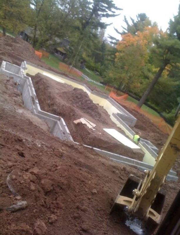 Construction site with excavated foundation walls, dirt, and trees in the background.