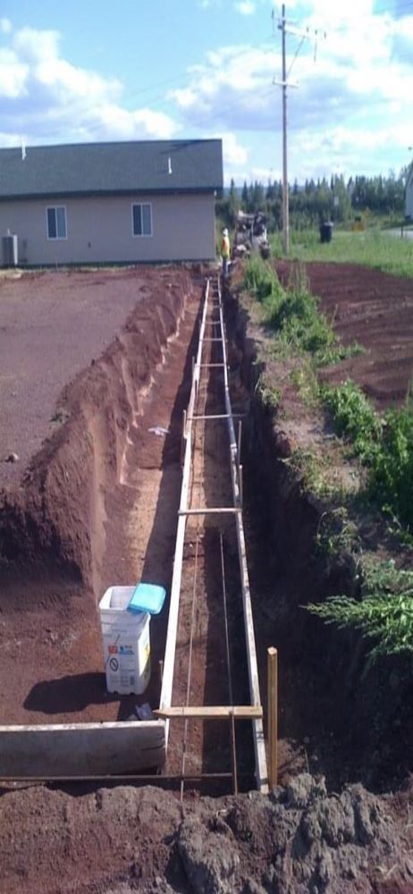 Construction site with a long trench, wooden forms, and a building in the background.