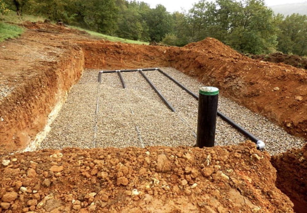 A trench with gravel and black pipes for a septic system, surrounded by dirt and trees.