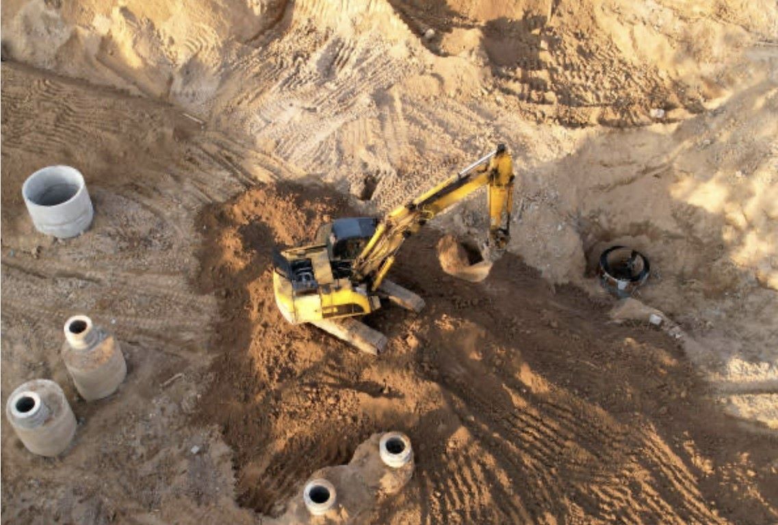 Yellow excavator digging in sandy soil at a construction site with concrete pipes.