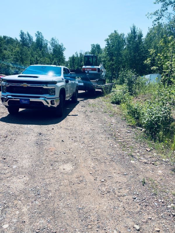 White pickup truck towing an excavator on a trailer on a gravel road. Sunny day, trees in the background.