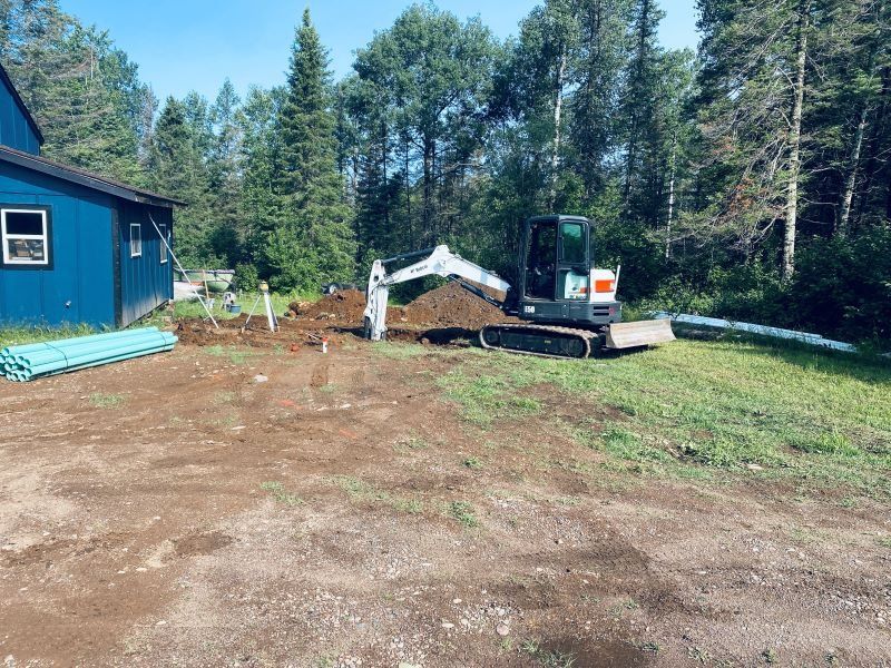 A small excavator digs near a blue building, surrounded by grass and trees. Construction site.