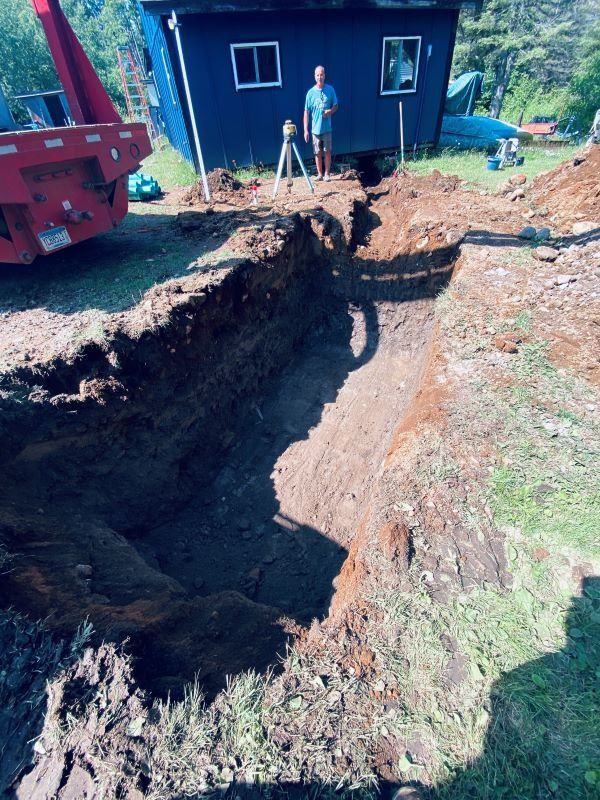 Man standing near trench, surveying, blue building in background, red container.