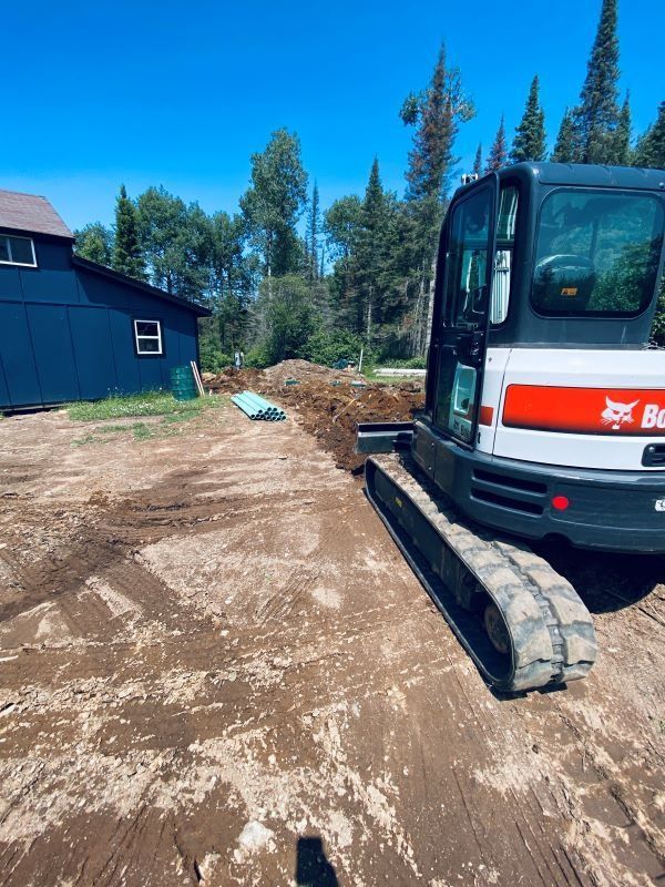A small excavator is digging a trench on a dirt lot next to a blue building, under a bright blue sky.
