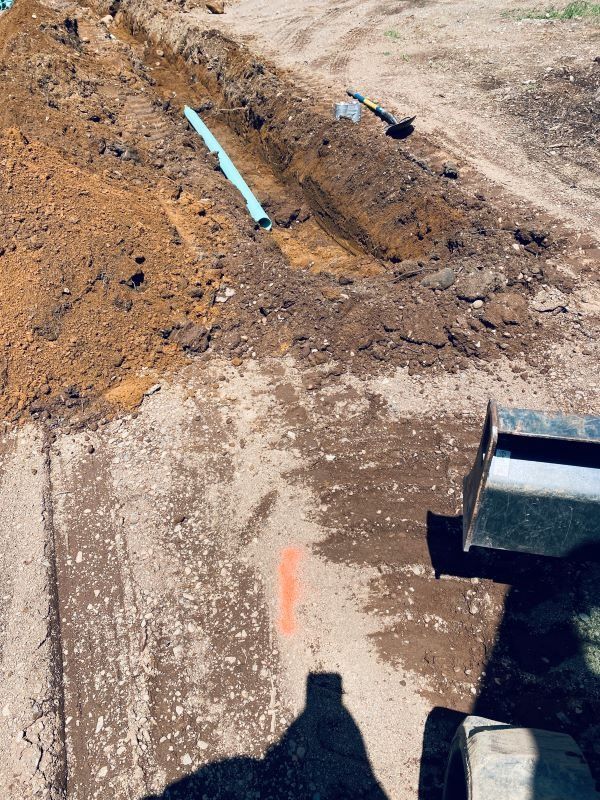 A trench with blue pipe, dug into the ground. A small excavator and orange marker are visible.