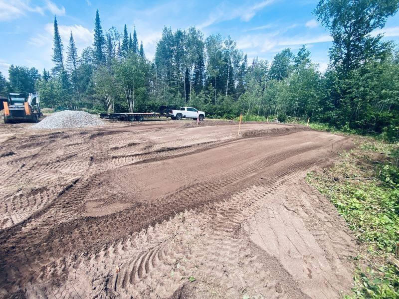 A dirt construction site, partially leveled, with a pickup truck and trees in the background.