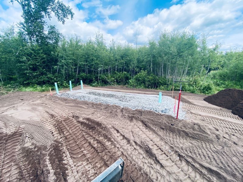 Construction site with gravel, pipes, and dirt, with trees in the background under a cloudy sky.