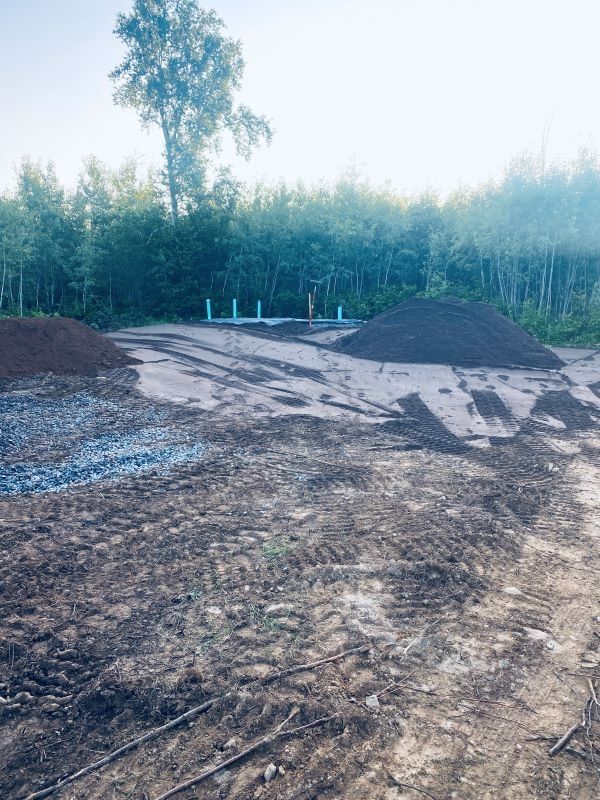 Construction site with dirt, gravel, and piles of soil, pipes in the middle, trees in the background.