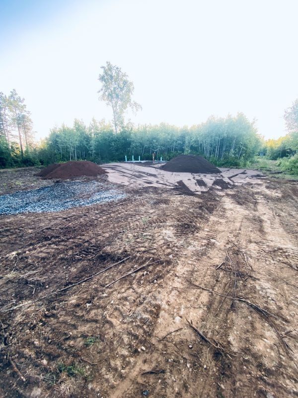 Dirt lot with piles of soil and gravel, surrounded by trees under a bright sky.