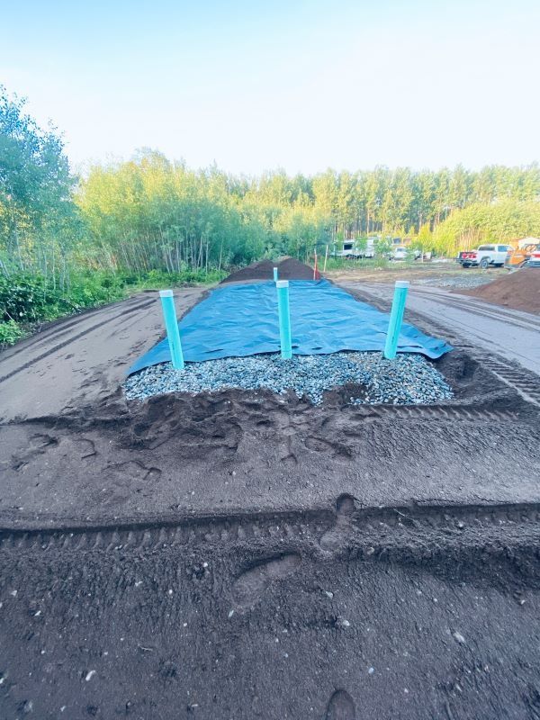Construction site: black fabric covered with gravel and pipes, dirt, and a treeline.