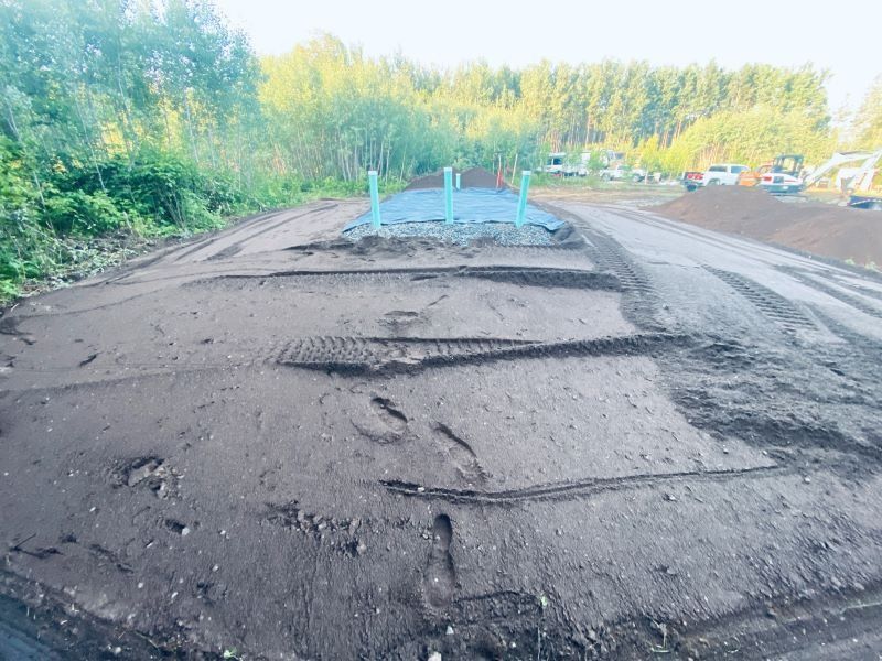 Dirt road construction site with blue pipes, trees, and equipment in the distance.