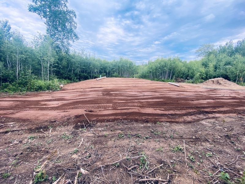 Cleared land area with reddish soil, surrounded by trees under a cloudy sky.