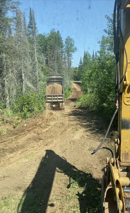Truck on a dirt road hauling material, trees line the sides, excavator arm in foreground.
