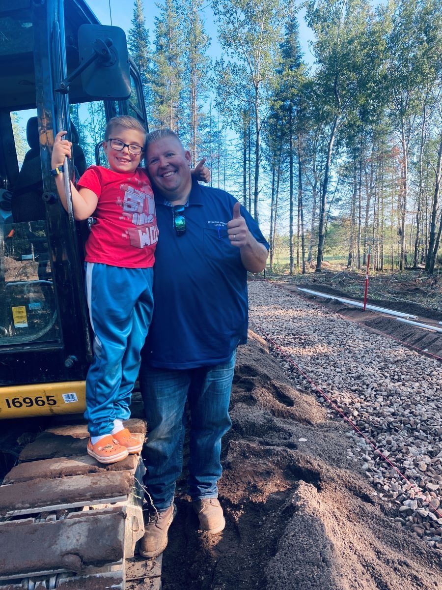 Man and child smiling, posing by construction equipment, thumbs up. Outdoors with gravel and trees.