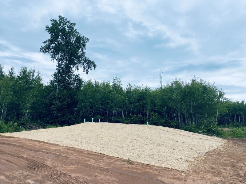 Gravel RV pad with utility posts, surrounded by trees under a cloudy sky.