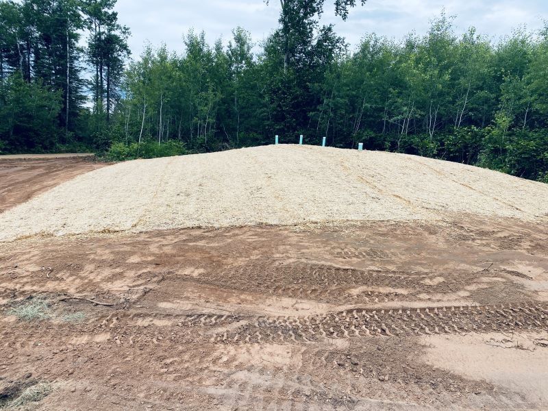 A large mound of light-colored material, with a sandy base, trees in the background.