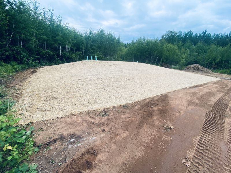 Gravel-covered construction site with pipes, surrounded by dirt and trees.
