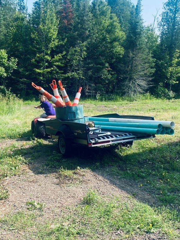 ATV carrying pipes and traffic cones on a trailer, in a grassy area with trees in the background.