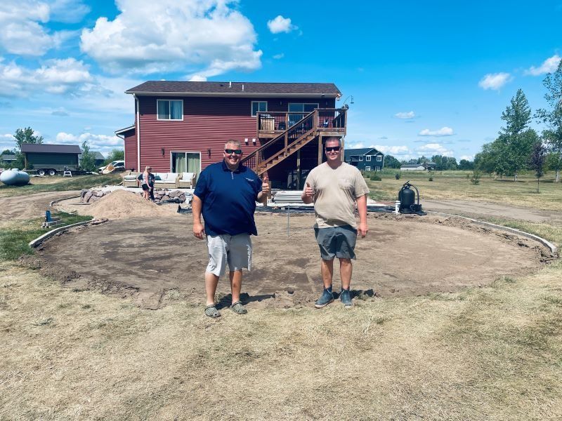 Two men stand in front of an excavation site for a pool, with a red house and blue sky in the background.