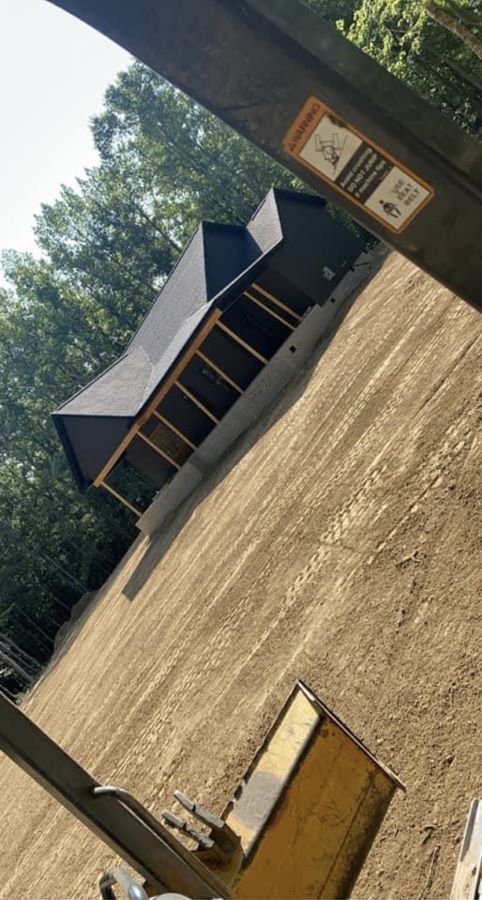 A house with a dark roof and porch sits on a graded dirt hill. Trees surround.